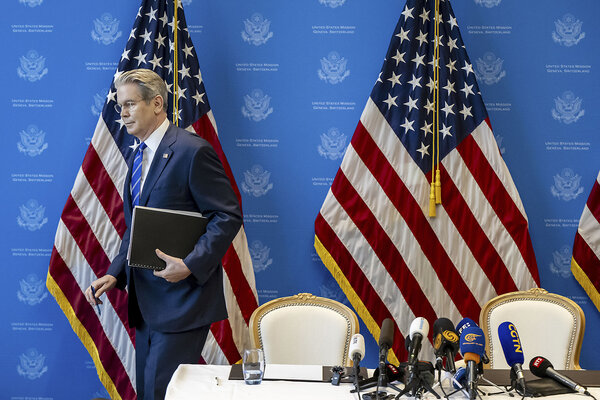 Scott Bessent steps away from a table with chairs and microphones, with American flags set up behind it.